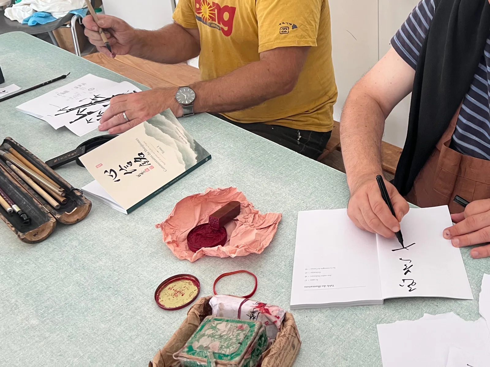 Two people at a table with calligraphy brushes, a wax seal and red ink, and a freshly printed copy of Commentaires du Shinjinmei, signing and stamping the finished book by hand